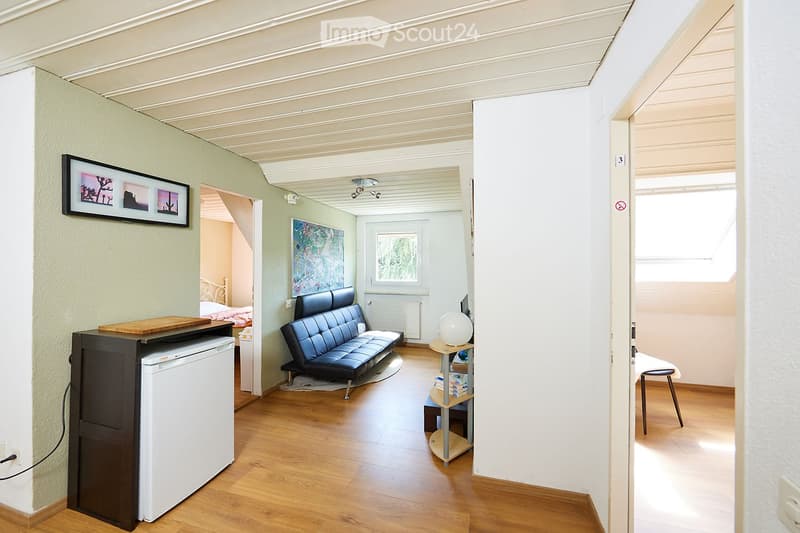 A modern, open-plan living and sleeping area with wooden flooring, a black leather sofa, a small table with a globe, a refrigerator, and a bed visible in the background.