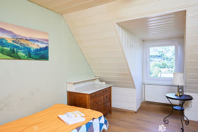 Cozy attic bedroom with wooden floors, slanted ceilings, a bed with orange and blue bedding, a wooden nightstand, a small round table with a lamp, and a painting on the wall.