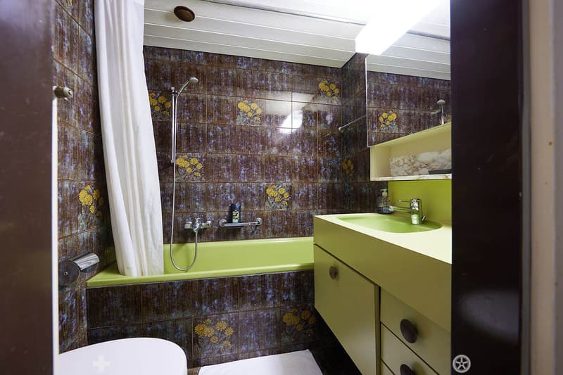 Bathroom with a green bathtub, white shower curtain, and a yellow sink vanity with a mirror and shelves. The walls are tiled with a floral pattern.