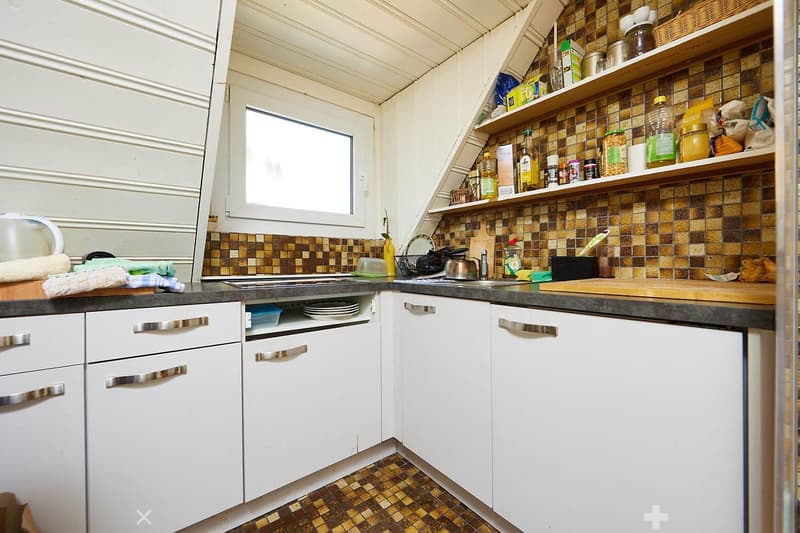 A compact kitchen with white cabinets, a dark countertop, and a tiled backsplash. The kitchen features a sink, a faucet, and various kitchen items on the countertop, including a cutting board, bottles, and cups.