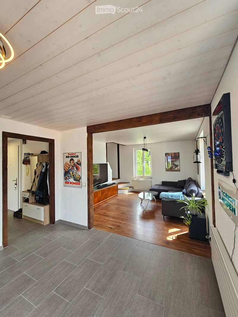 Modern living room with wooden floors, white walls, and exposed wooden beams. Features a grey sofa, coffee table, TV, cabinet, and a plant on a side table.