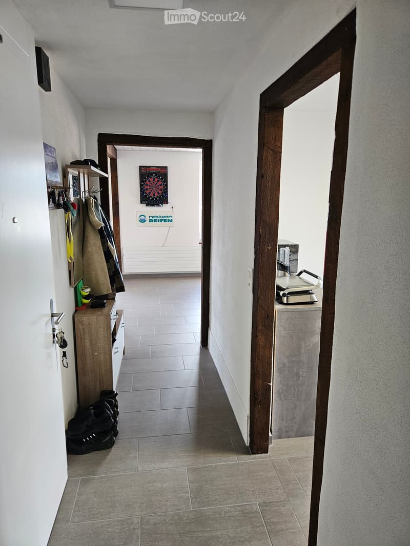 A narrow hallway with tiled flooring, white walls, and wooden beams. Visible items include a coat rack with coats and shoes, a small wooden cabinet, a dartboard, and a small kitchen area with a blender and other appliances.