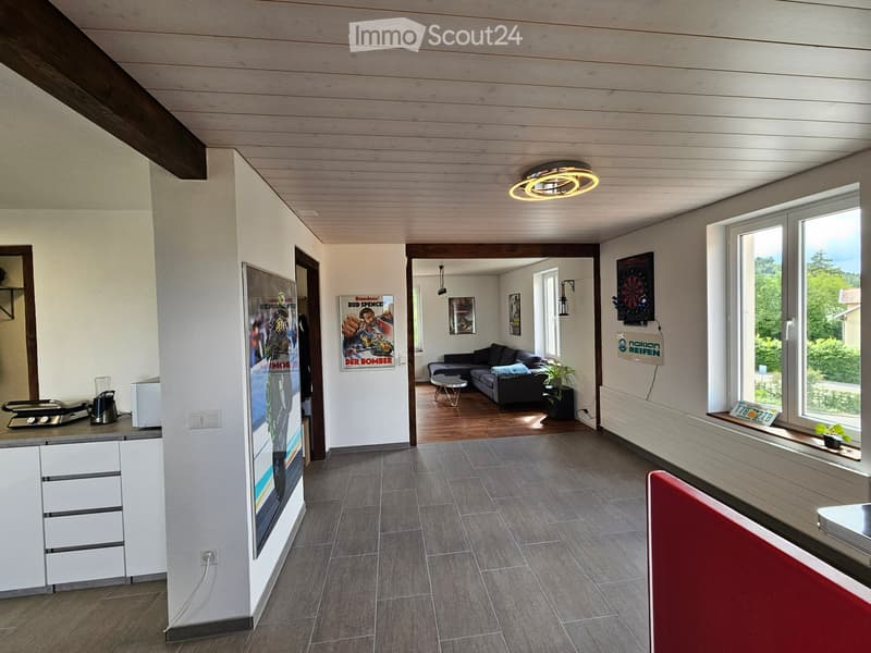 Open-plan living and kitchen area with grey sofa, coffee table, kitchen cabinet, blender, fridge, posters, dartboard, and a view of the garden through large windows.