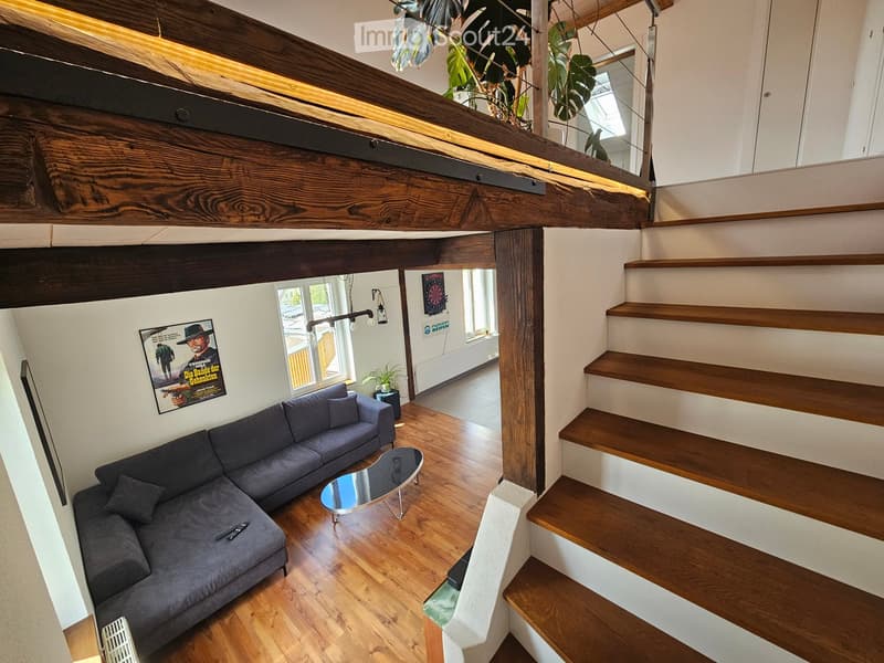 Living room with a grey sectional sofa, a round glass coffee table, a remote control on the sofa, a poster on the wall, a small plant, and a table lamp. Wooden staircase leads to an upper level.
