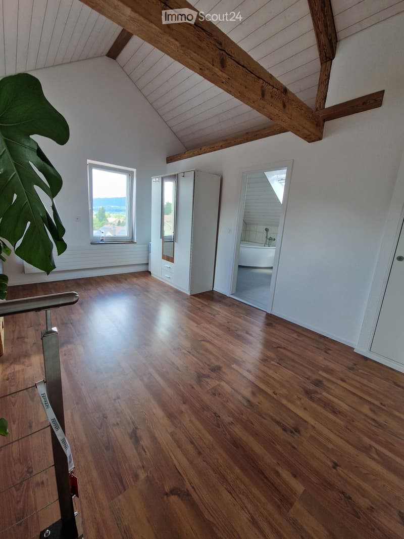 Empty room with wooden flooring, white walls, exposed wooden beams on the ceiling, a large window with a view, and an open doorway leading to a bathroom.