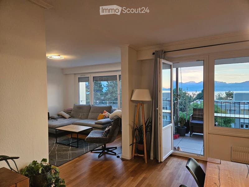 Modern living room with wooden flooring, large windows, and a balcony offering a scenic view. Features a grey sofa, coffee table, armchair, floor lamp, and a small table with a chair.