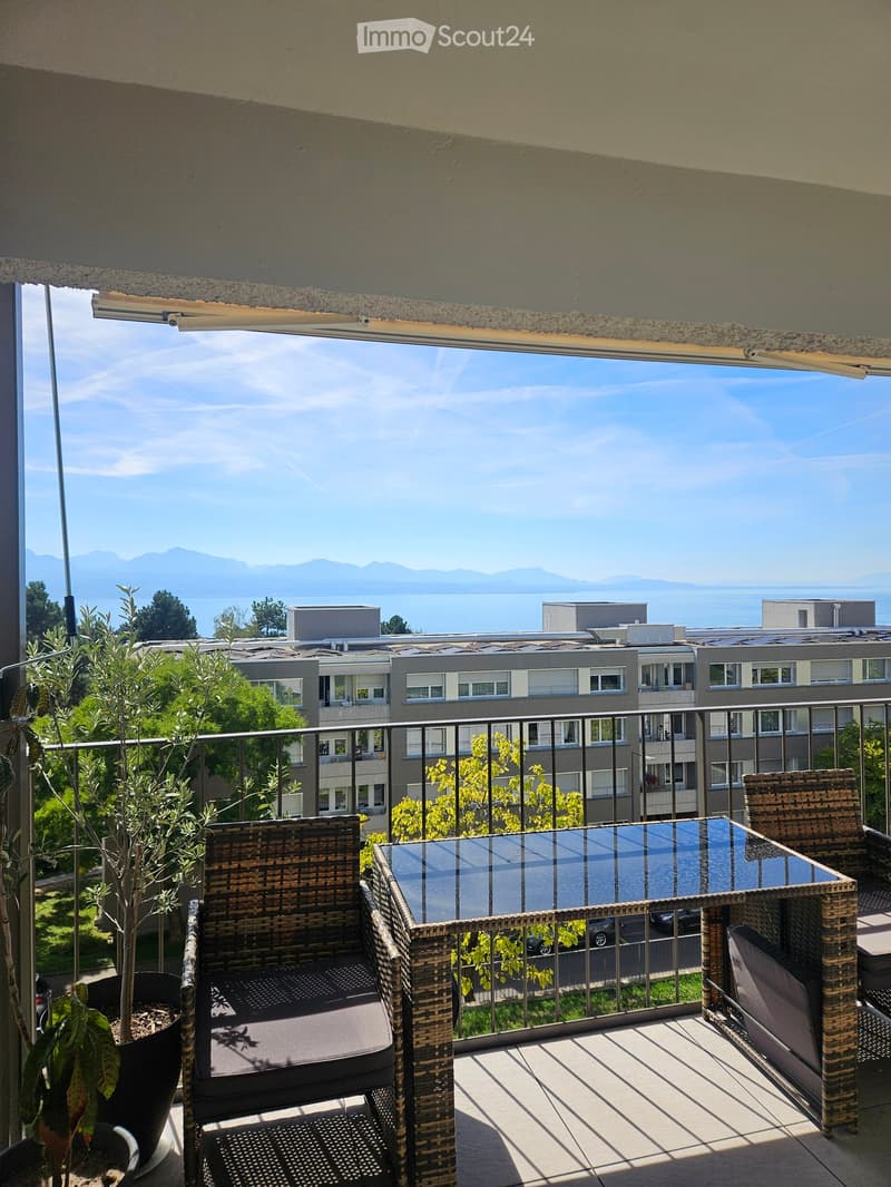 Balcony with wicker chairs, a glass table, and a potted plant, overlooking a scenic view of a lake and mountains.
