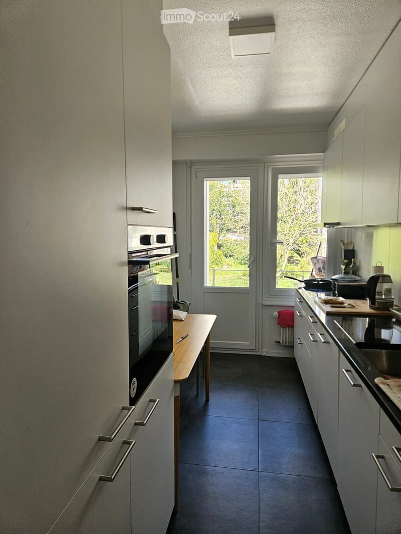 Modern kitchen with grey cabinets, dark tiled floor, and a wooden table near the oven. The kitchen features a sink, cutting board, and various appliances including a coffee maker and a pot on the counter.