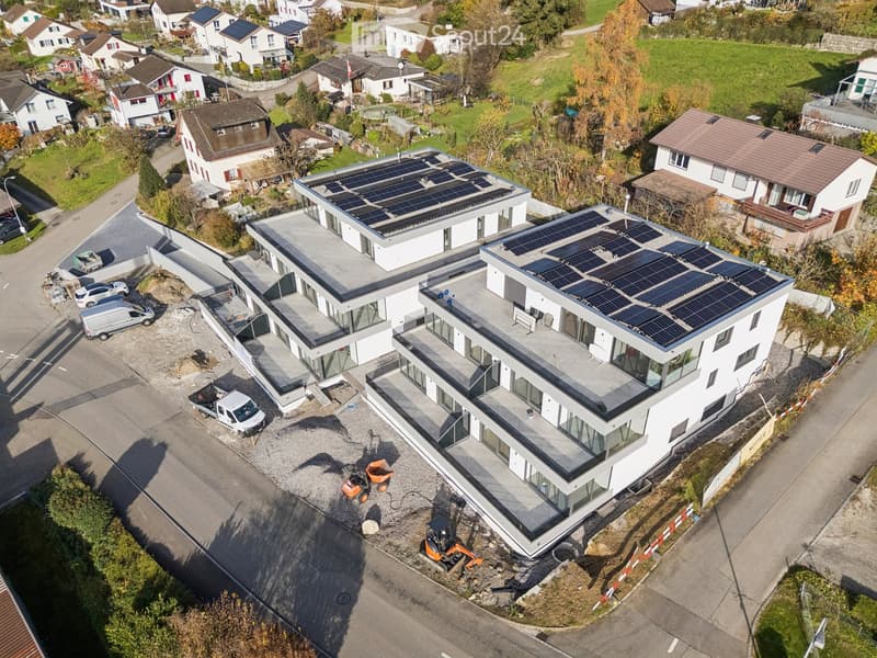 Multi-story apartment building with solar panels on the roofs, glass facades, surrounded by residential homes, green fields, and a road.