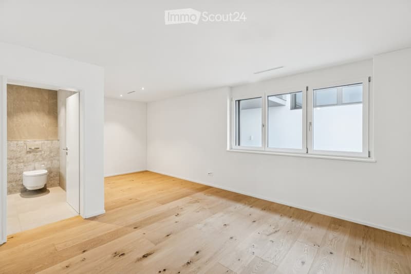 Empty room with hardwood floors, white walls, large window, small doorway leading to bathroom
