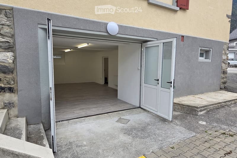 Open garage with wooden floor and white walls, adjacent to a stone building with a small window and a satellite dish.
