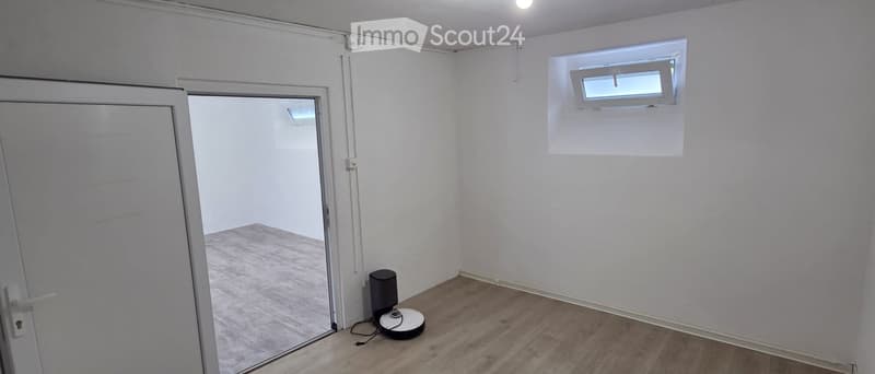 Empty room with white walls, wooden flooring, and a small window. A robotic vacuum cleaner is visible on the floor.