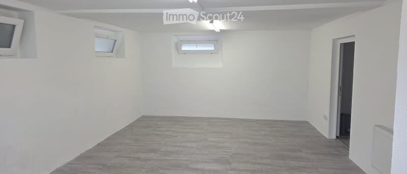 Empty room with white walls, grey tiled floor, and three windows, one of which is a skylight. There is a door leading to another space.