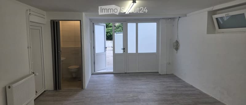 Empty hallway with wooden flooring, white walls, and a radiator on the left side. An open door leads to a small bathroom with a toilet and sink, and a large sliding glass door provides access to the outdoors.