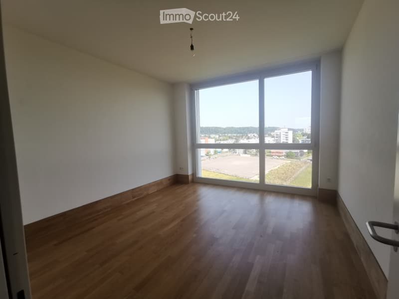 Empty room with wooden flooring, white walls, and large windows offering a view of the outside area.