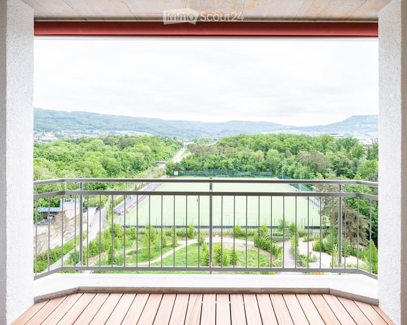 Balcony with wooden decking, metal railing, overlooking a green landscape with trees, a sports field, and distant hills.