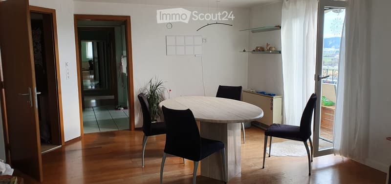 Dining room with a round wooden table, four chairs, a small plant, and a shelf with decorative items. The room has wooden flooring and large windows with white curtains.