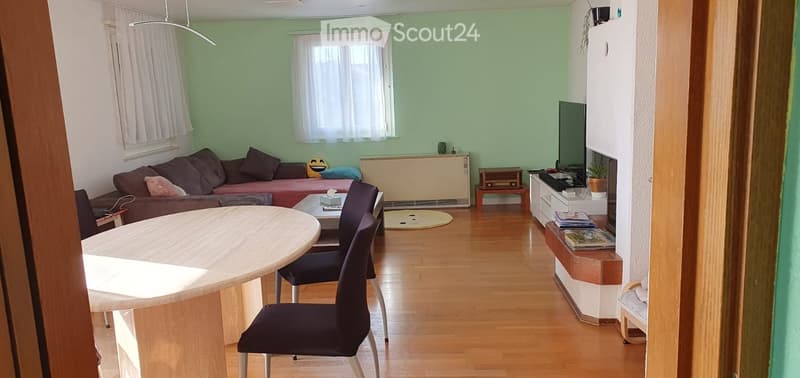A cozy living room with a sofa, a dining table with chairs, a television, a small side table, and a plant. The room features light green walls, wooden flooring, and large windows with white curtains.