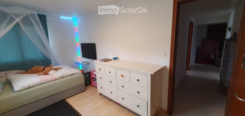 A well-lit bedroom with a bed, a dresser, a small stool, a screen, and a toy on the bed.