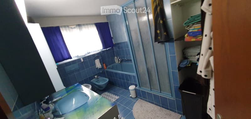 Bathroom with blue tiles, a sink, bathtub, mirror, towel rack, and shelves with folded towels and a bathrobe.