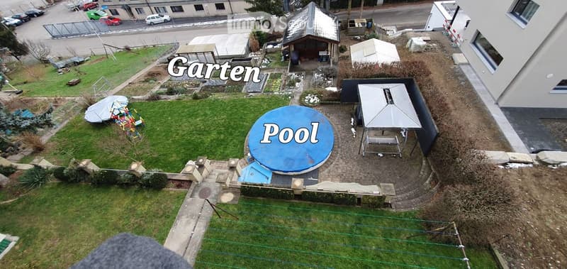 Aerial view of a garden area with a round swimming pool covered by a blue tarp, surrounded by a paved patio and grassy lawn.