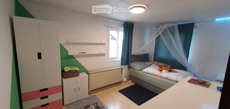 A small, well-lit bedroom with a bed, wardrobe, shelves, and a table. The room features a window with curtains, a radiator, and a colorful accent wall.