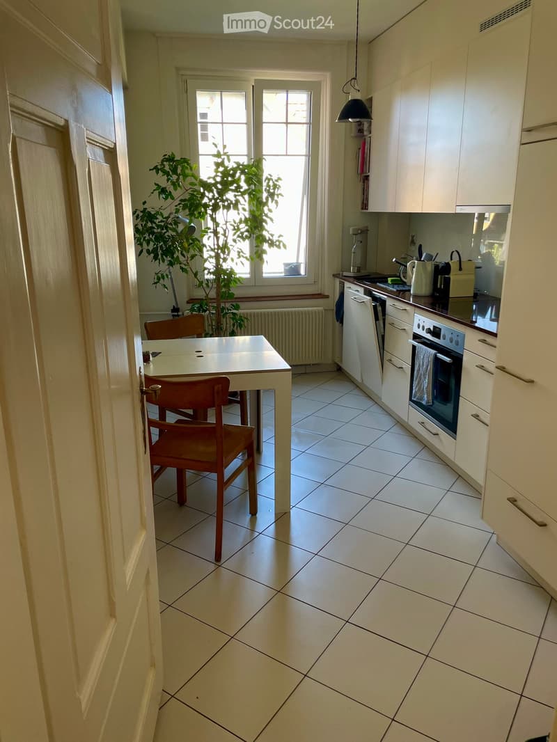 Modern kitchen with white tiled floor, white cabinets, a small dining table with two chairs, and various appliances including an oven and coffee maker. A large window with a potted plant is visible.