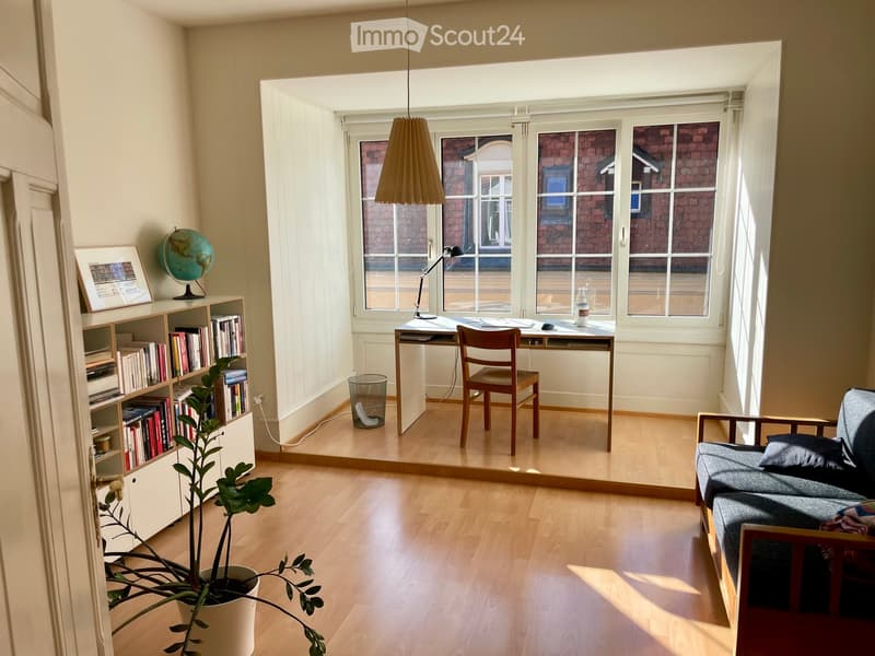 Bright, open living space with wooden flooring, large windows, a sofa, a desk with a chair, a bookshelf, a globe, and a potted plant.