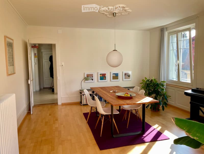 Bright dining area with wooden floor, large wooden dining table, white chairs, bowl of fruit, pendant light, plant, and picture frames on the wall.