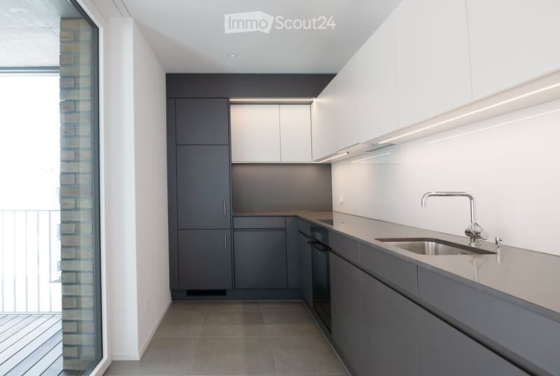 Modern kitchen with white and grey cabinets, a stainless steel sink, and a sleek faucet. The space features a large window with a view of a brick wall and balcony.