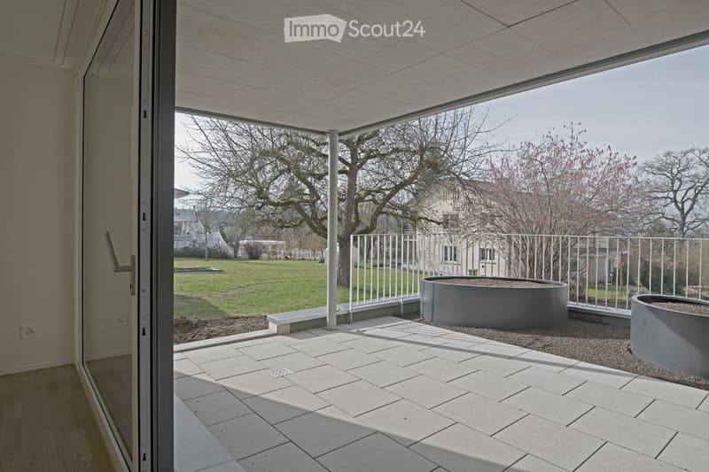 Modern balcony with tiled floor, white railing, and two grey planters, overlooking a park with trees and open space.