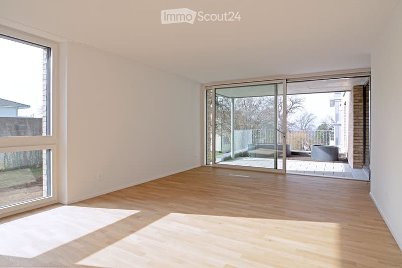 Bright, empty room with light wooden flooring and large windows, featuring a sliding glass door leading to a patio area.