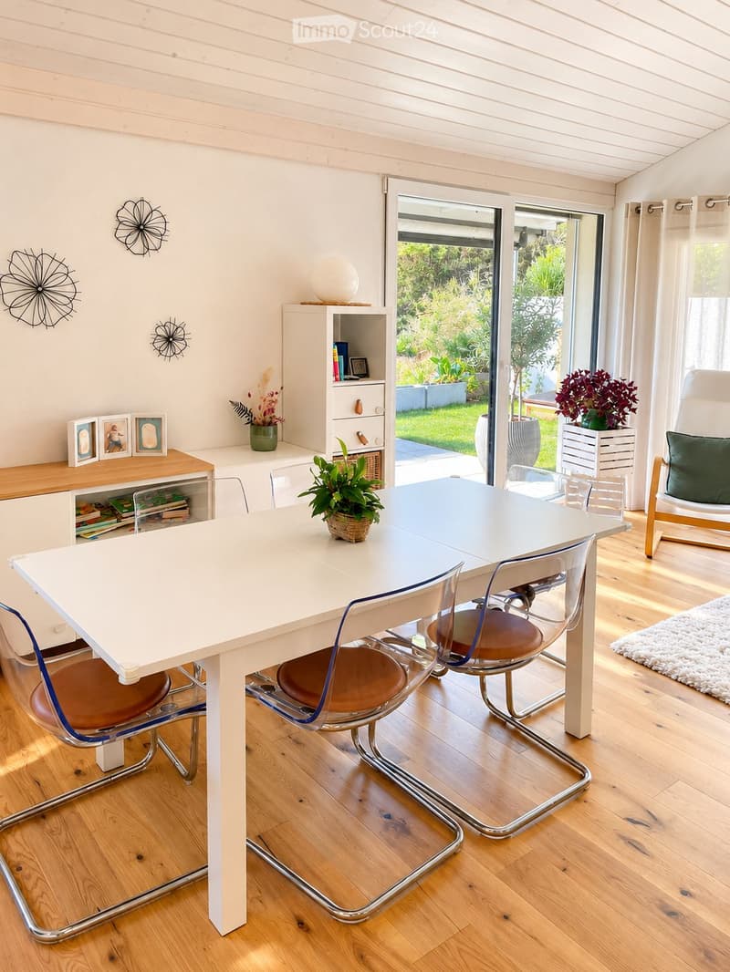 Bright dining area with a white table, modern chairs, a cabinet, and a view to the garden through large glass doors.