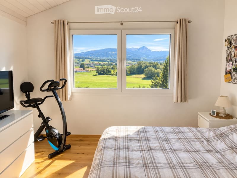 Bright bedroom with wooden flooring, large windows offering scenic mountain views, a neatly made bed with a plaid blanket, an exercise bike, a nightstand with a lamp, and a TV on a white cabinet.