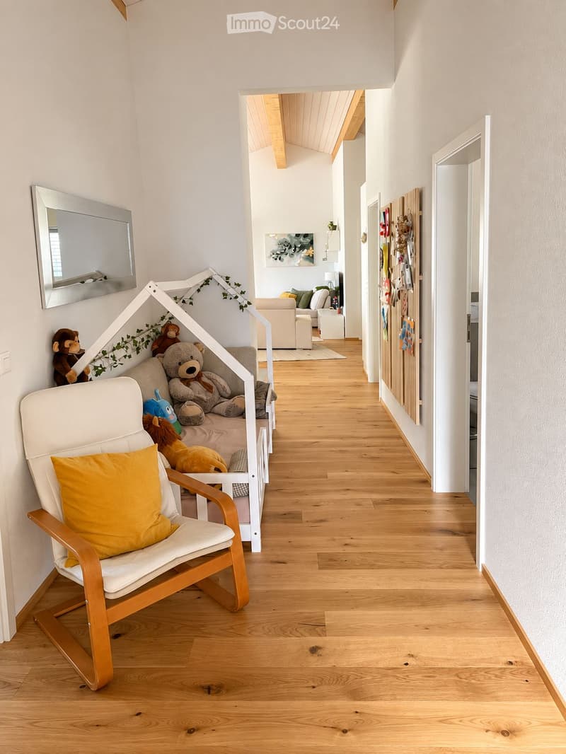 Bright hallway with wooden flooring, white walls, and a small seating area with a rocking chair and stuffed animals. A mirror is mounted on the wall, and a living room is visible at the end of the hallway.