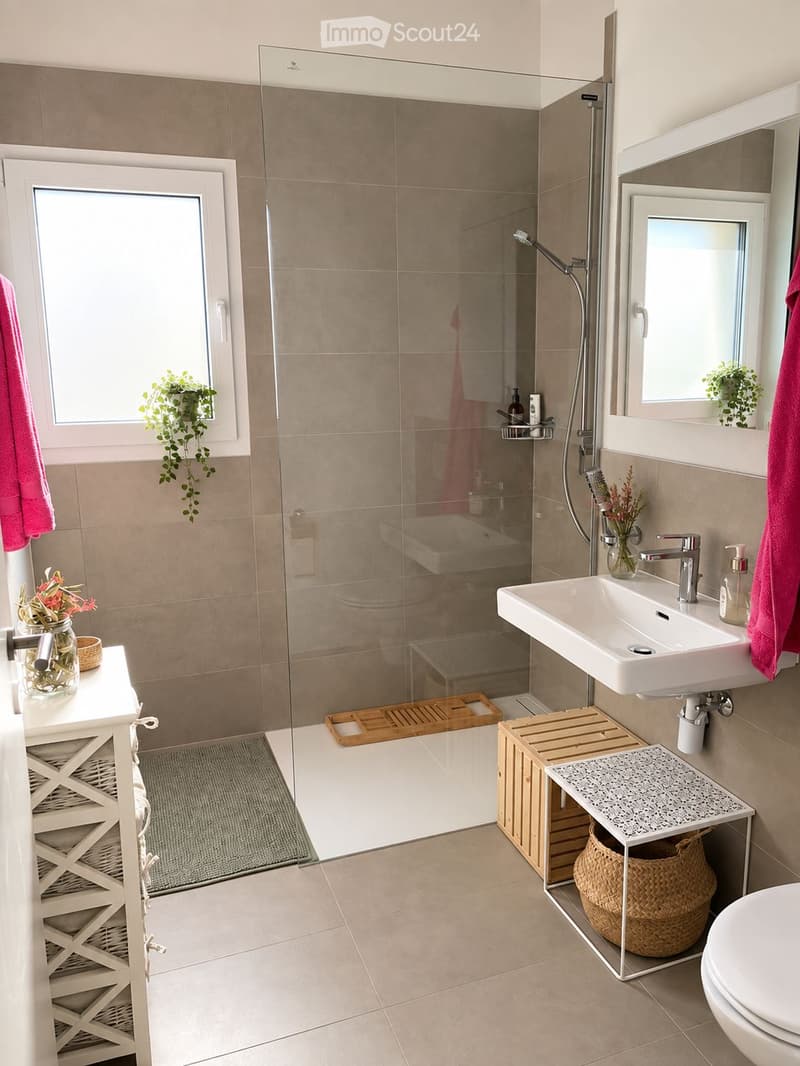 Modern bathroom with a glass-enclosed shower, white sink, mirror, and a small wooden shelf holding a basket. The space features beige tiles, a grey rug, and decorative plants.