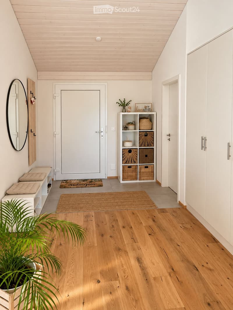 Bright hallway with wooden flooring, white walls, and a slanted ceiling. Features include a large mirror, a small shelf with decorative items, a potted plant, and a white bench with cushions.