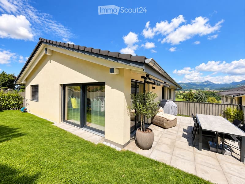 Modern house with a well-maintained garden and a patio area featuring a table, chairs, a wicker sofa, and a potted plant. The house has large glass doors and a clean, contemporary design.