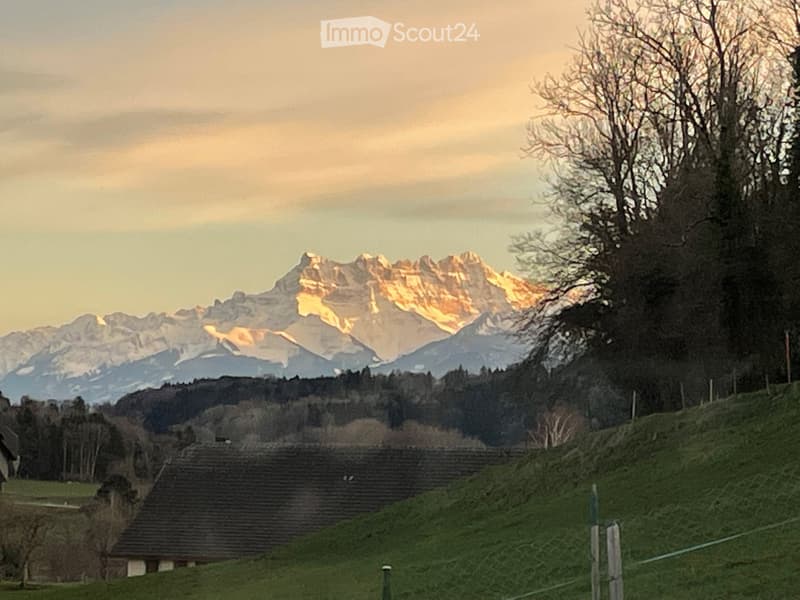 A serene outdoor scene featuring a grassy hillside, a small house, and a stunning mountain range with snow-capped peaks in the background.