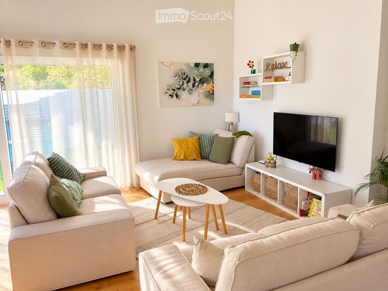Modern living room with light-colored furniture, a large window with sheer curtains, a television mounted on the wall, and decorative shelves.