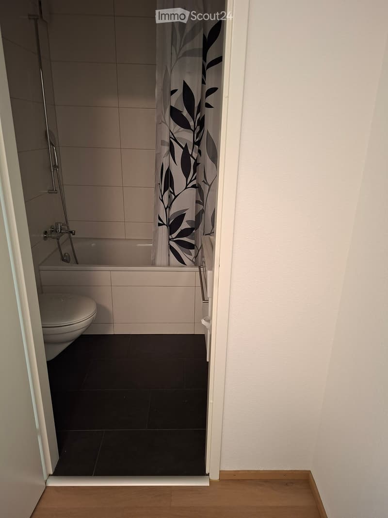 Modern bathroom with a toilet, bathtub, and shower curtain featuring leaf patterns. The floor is tiled in dark black tiles, and the walls are tiled in light beige tiles.