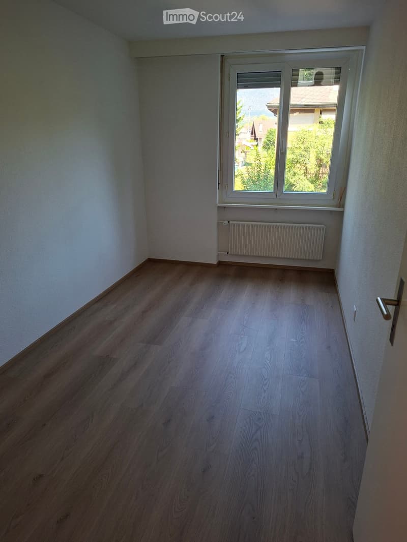 Empty room with wooden flooring, white walls, a window with white frame, and a radiator below the window.