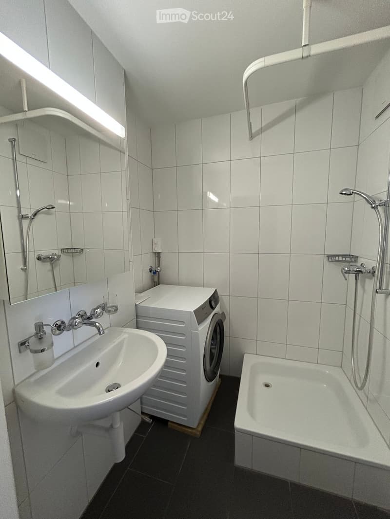 Modern bathroom with white tiles, a sink, bathtub, and a compact washing machine. Features include a showerhead, faucet, and a mirror above the sink.
