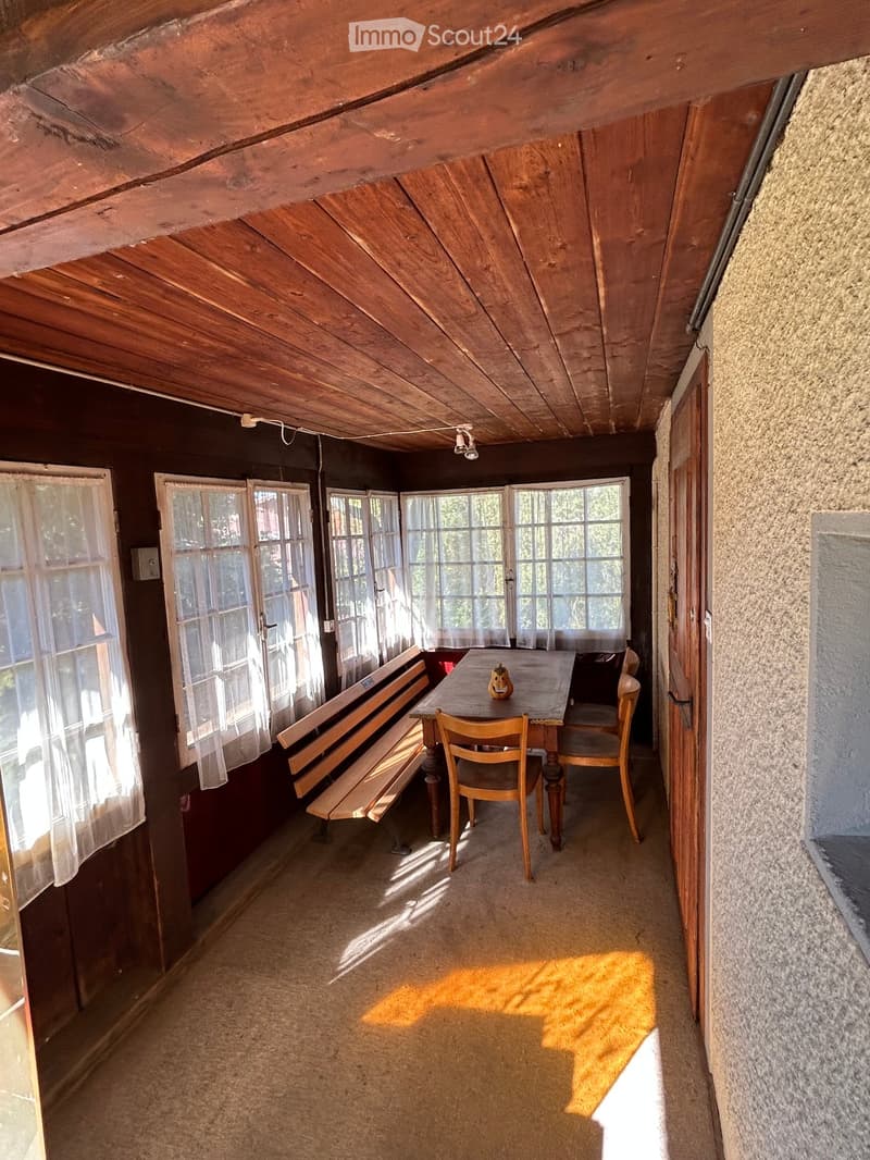 A rustic wooden room with exposed wooden beams, large windows with sheer curtains, a wooden table with chairs, and a bench along the wall.