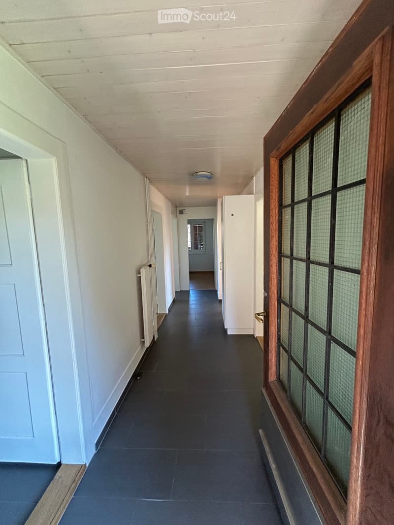 A long, narrow hallway with dark blue tiled flooring, white walls, and several doors on both sides. The ceiling has a simple white finish with a single light fixture. A radiator is visible on the left wall, and a wooden door with frosted glass panels is o