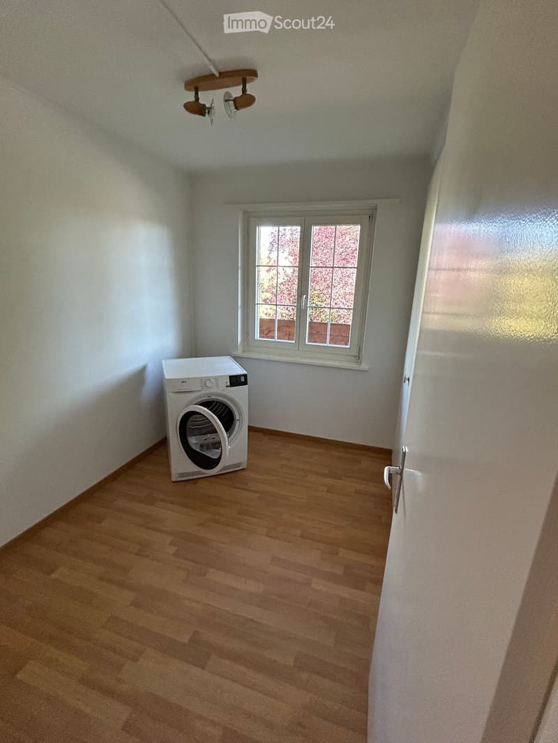 A small room with wooden flooring, white walls, and a single window. A white washing machine is positioned near the window.