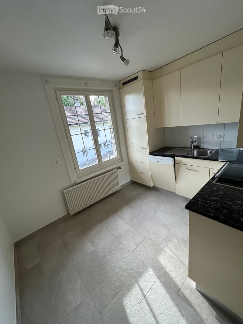 Empty kitchen with light-colored cabinets, a sink, a dishwasher, and a window overlooking a neighboring building.