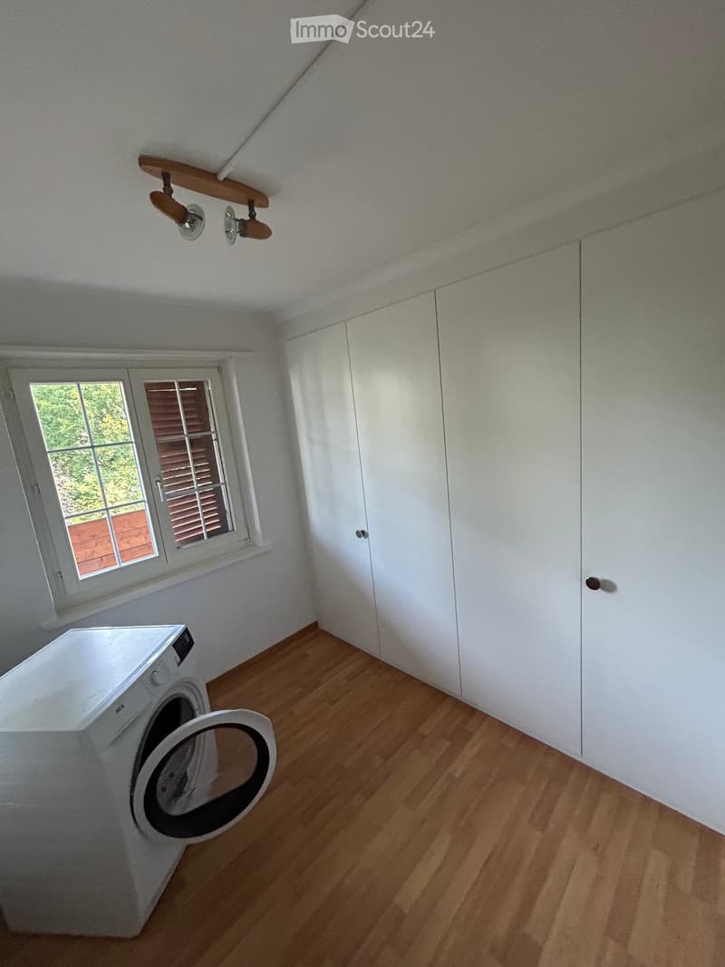 A small, well-lit room with wooden flooring, a white washing machine, and built-in white wardrobes. The room has a slanted ceiling and a window with blinds.