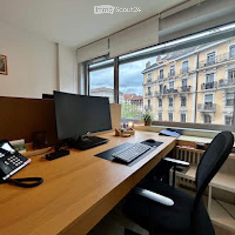 Office space with a wooden desk, computer, chair, telephone, and radiator near a large window overlooking a cityscape.