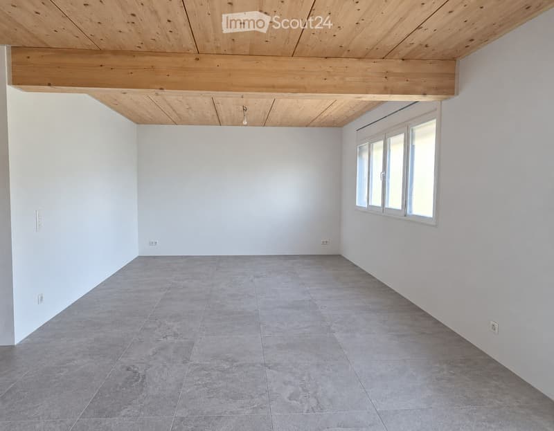 Empty room with white walls, grey tiled floor, and a large window with multiple panes. The ceiling features exposed wooden beams.